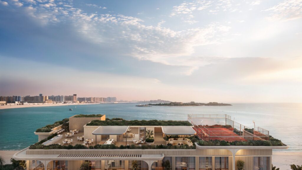 Soma Residences est un immeuble moderne situé en bord de mer, doté d'un toit-terrasse végétalisé, d'espaces de restauration en plein air et d'un court de tennis rouge donnant sur une mer calme et une plage de sable, sous un ciel partiellement nuageux, avec au loin la silhouette de la ville qui se profile le long du littoral.