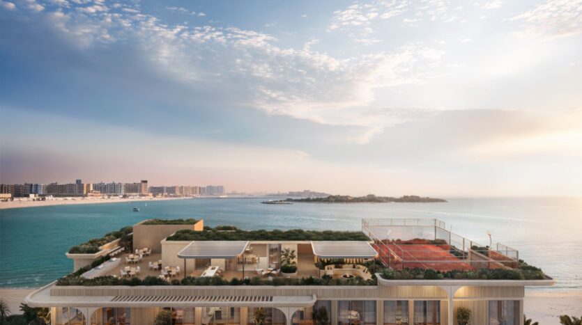 Soma Residences est un immeuble moderne situé en bord de mer, doté d'un toit-terrasse végétalisé, d'espaces de restauration en plein air et d'un court de tennis rouge donnant sur une mer calme et une plage de sable, sous un ciel partiellement nuageux, avec au loin la silhouette de la ville qui se profile le long du littoral.