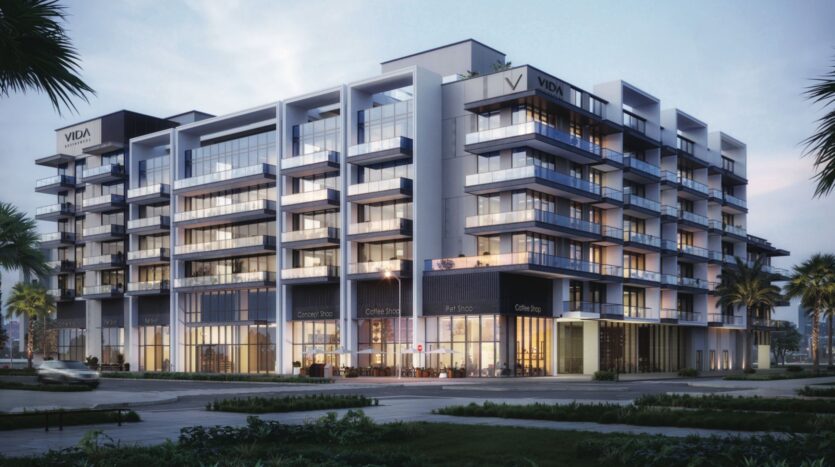 Un immeuble moderne de plusieurs étages avec de grandes baies vitrées et des balcons, avec des boutiques au rez-de-chaussée et l'enseigne "VIDA" dans les coins supérieurs - Vida Residences Saadiyat Island - entouré de palmiers et d'une rue tranquille.