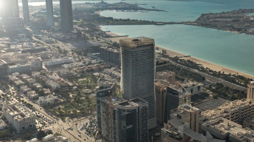 Vue aérienne d'une ville côtière où se trouve Vida Residences Saadiyat Island, avec des immeubles de grande hauteur, des routes et des espaces verts au bord d'une mer turquoise. En arrière-plan, des plages et une étendue de terre s'avancent dans l'eau.