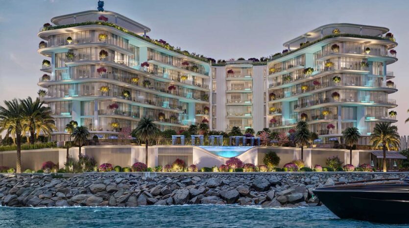 La luxueuse et moderne Villa Del Garda, située en bord de mer, se compose de deux ailes incurvées, de balcons en verre, de palmiers, de fleurs colorées et d'une piscine centrale éclairée. Elle est vue de l'eau, avec une digue en pierre devant et un bateau partiellement visible sur la droite.
