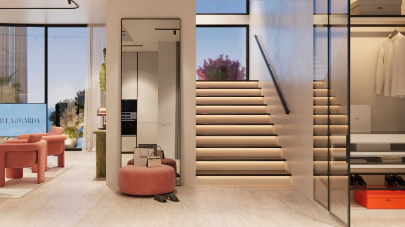 Un espace de vie moderne et minimaliste à Villa Del Garda avec des chaises et un ottoman roses, un grand miroir, un placard ouvert et un escalier lumineux avec éclairage intégré. De grandes fenêtres inondent la pièce de lumière naturelle.