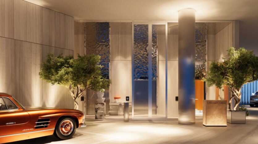Un hall d'entrée luxueux et moderne avec de hautes portes vitrées, des arbres verdoyants, un éclairage chaleureux et une voiture vintage brillante de couleur cuivre garée à l'intérieur. Un petit bureau de réception indique "VILLA ARTEFACTO" à la Villa Del Garda.