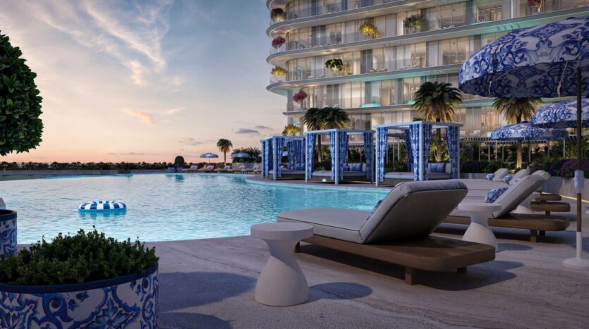 Piscine luxueuse au coucher du soleil à la Villa del Garda, avec des chaises longues rembourrées, des parasols bleus et blancs, des cabanas, des palmiers et un bâtiment moderne incurvé. La piscine tranquille reflète le ciel tandis que des plantes décoratives bordent la scène sereine.