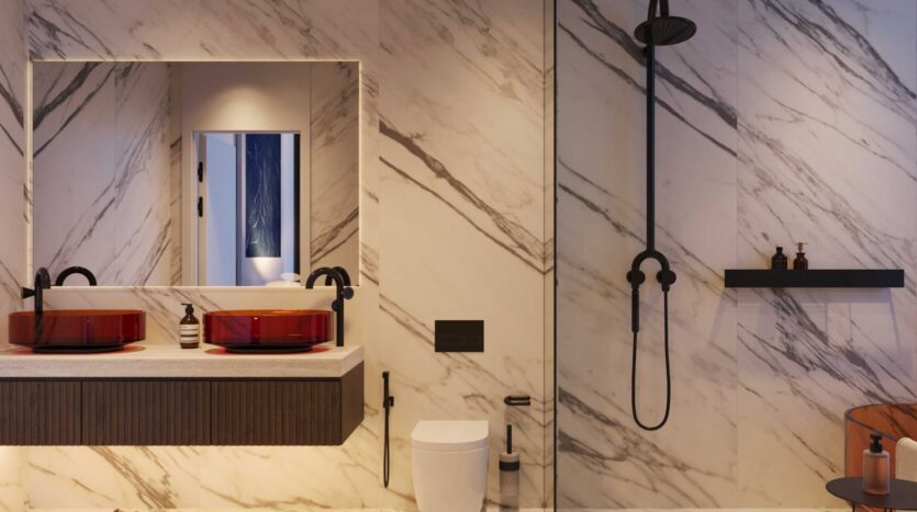 Salle de bains moderne de la Villa Del Garda avec des murs en marbre blanc, un meuble à double vasque avec des lavabos en verre rouge et des accessoires noirs, des toilettes murales, une pomme de douche noire et une étagère contenant des bouteilles brunes. Un miroir reflète l'ouverture de la porte.