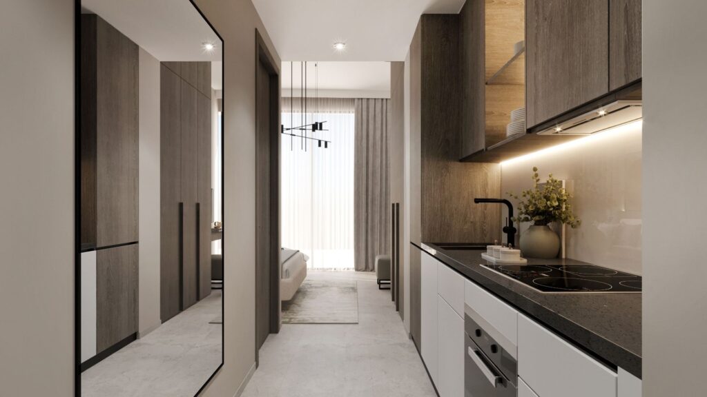 Une cuisine moderne et épurée, avec des meubles en bois clair et foncé, des plans de travail noirs, une cuisinière encastrée et un éclairage sous les meubles — idéale pour ceux qui souhaitent investir dans un studio à Dubaï. Un vase de fleurs trône près de l'évier ; le couloir mène à une chambre dotée de grandes fenêtres.