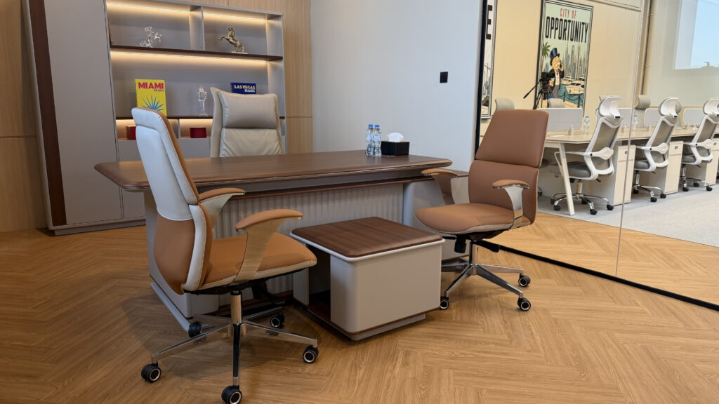 Un bureau moderne équipé d'un bureau en bois, de deux chaises marron et blanches et d'une petite table. Des étagères remplies de livres et une affiche sur laquelle on peut lire « OPPORTUNITY » ajoutent du charme à l'ensemble, tandis que la baie vitrée dévoile d'autres bureaux à Dubaï destinés aux investisseurs chez Dubai Immo.