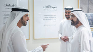 Trois hommes vêtus de kanduras blanches traditionnelles et portant un couvre-chef se tiennent à l'intérieur, en pleine conversation et souriants, avec en arrière-plan des affiches d'information « Dubai-it » et une « nouvelle expression » accrochées au mur.