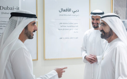 Trois hommes vêtus de kanduras blanches traditionnelles et portant un couvre-chef se tiennent à l'intérieur, en pleine conversation et souriants, avec en arrière-plan des affiches d'information « Dubai-it » et une « nouvelle expression » accrochées au mur.