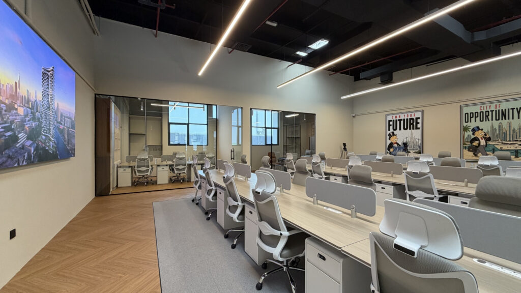 Un espace de bureaux moderne avec des rangées de bureaux et de chaises vides, de grandes fenêtres, une salle de réunion aux parois vitrées et des affiches accrochées aux murs. Un écran numérique « Dubai Immo » diffuse une vue panoramique de la ville. Le bureau est bien éclairé et bien agencé : l'endroit idéal pour les investisseurs à la recherche de bureaux à Dubaï.