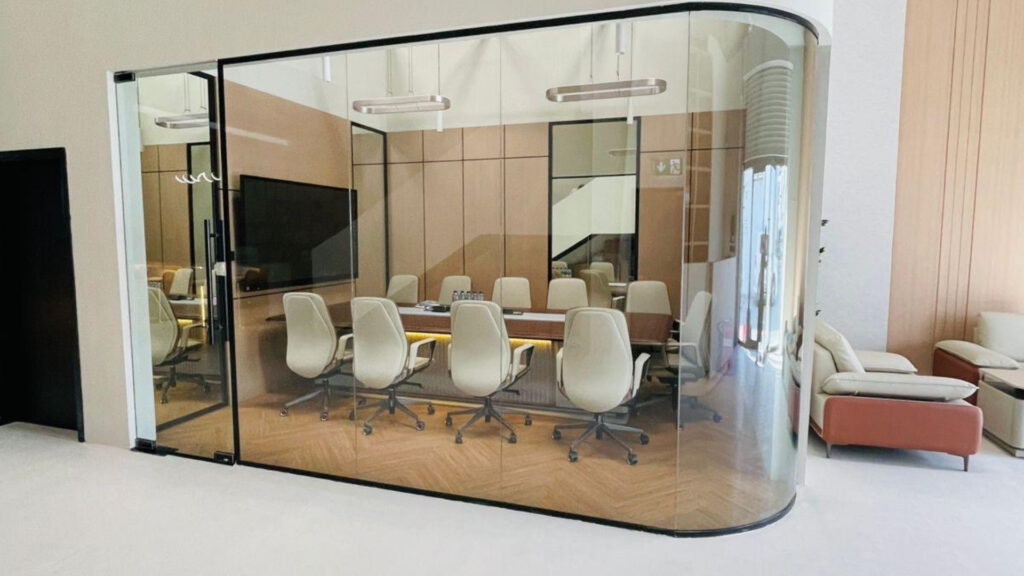 Une salle de réunion moderne aux parois vitrées, dotée d'une grande table en bois entourée de huit chaises de bureau blanches, idéale pour les investisseurs. Cet espace bien éclairé, proposé par Dubai Immo, jouxte un coin salon et met en valeur des bureaux haut de gamme dans le style de Dubaï.