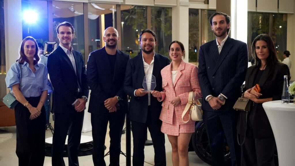 Un groupe de sept personnes, vêtues de tenues professionnelles et semi-formelles, se tient à l'intérieur lors d'un événement bien éclairé. Au centre, un homme tient un trophée en verre transparent récompensant le « TOP BROKER » des Mansory Residences, organisé par Dubai Immo. On aperçoit des tables modernes à l'arrière-plan.