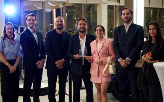 Un groupe de sept personnes, vêtues de tenues professionnelles et semi-formelles, se tient à l'intérieur lors d'un événement bien éclairé. Au centre, un homme tient un trophée en verre transparent récompensant le « TOP BROKER » des Mansory Residences, organisé par Dubai Immo. On aperçoit des tables modernes à l'arrière-plan.