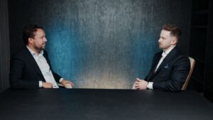 Deux hommes en costume sont assis face à face à une table noire, en pleine conversation au sujet des rendements à Dubaï. L'arrière-plan est sombre, avec une teinte bleutée, et l'éclairage est concentré sur les deux hommes.
