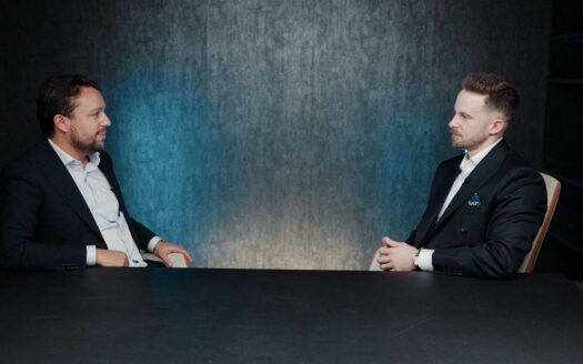 Deux hommes en costume sont assis face à face à une table noire, en pleine conversation au sujet des rendements à Dubaï. L'arrière-plan est sombre, avec une teinte bleutée, et l'éclairage est concentré sur les deux hommes.