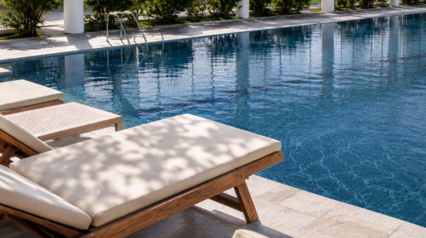 Une chaise longue en bois « Beverly Crown » de la marque HMB, dotée d'un coussin beige, trône au bord d'une piscine d'un bleu limpide, entourée de verdure et de colonnes blanches. Les rayons du soleil projettent des ombres tachetées sur la terrasse de la piscine.