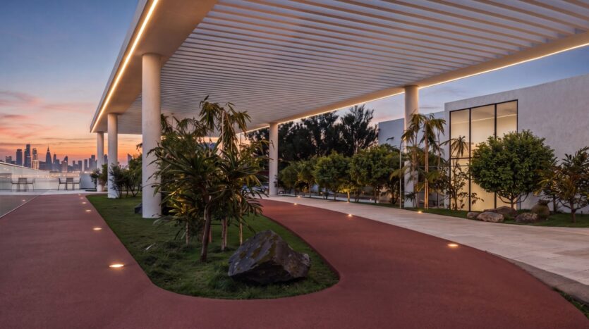 Une allée extérieure moderne du complexe Beverly Crown, conçu par HMB, se compose d'un chemin rouge sous une pergola blanche, bordée de plantes et d'arbres luxuriants, éclairée par des lampes encastrées dans le sol au coucher du soleil, avec la silhouette de la ville en arrière-plan.