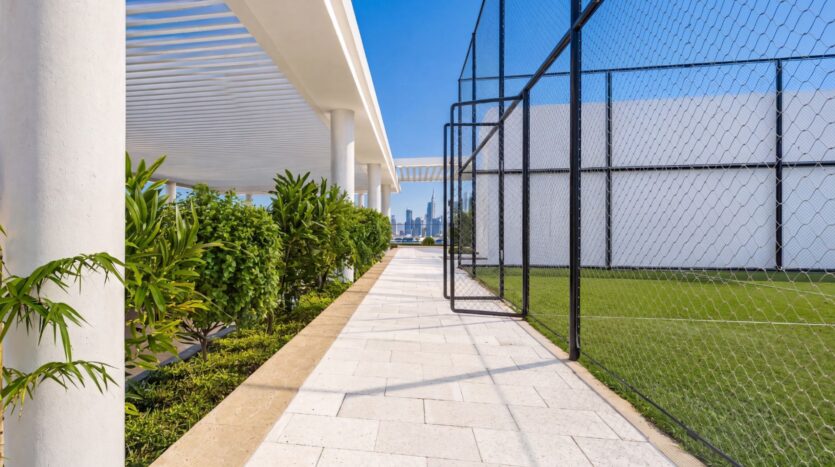 Une allée en plein air baignée de soleil au sein du complexe Beverly Crown by HMB, bordée d'un côté par des plantes vertes et de l'autre par un terrain de sport clôturé, avec la silhouette de la ville visible en arrière-plan sous un ciel bleu limpide.