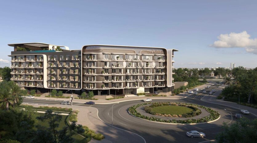 Un hôtel moderne de plusieurs étages doté de balcons, baptisé Zoya, se dresse à côté d'un rond-point entouré d'espaces verts aménagés, tandis que plusieurs voitures circulent sur les routes adjacentes sous un ciel dégagé. Ce cadre animé fait partie du complexe « Mirari Lagoon by Zoya ».