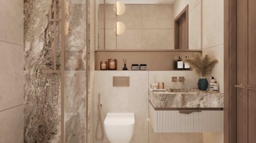 Salle de bains moderne équipée d'un WC suspendu, de plans de travail en marbre, d'un meuble-lavabo suspendu, d'une douche vitrée, de robinetterie dorée, de pots décoratifs et d'herbe de la pampa dans un vase. Les touches de « Mirari Lagoon » de Zoya apportent une note subtile de couleur apaisante aux tons neutres, tandis qu'un éclairage tamisé crée une atmosphère chaleureuse et élégante.