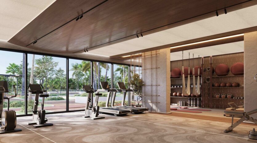 Une salle de sport moderne équipée d'appareils de cardio face à de grandes baies vitrées, d'anneaux de gymnastique, de ballons médicinaux et de divers équipements de fitness soigneusement rangés : un véritable havre de bien-être au Mirari Lagoon by Zoya. À l'extérieur, les palmiers et la végétation sont visibles à travers les baies vitrées, offrant une vue apaisante pendant votre entraînement.