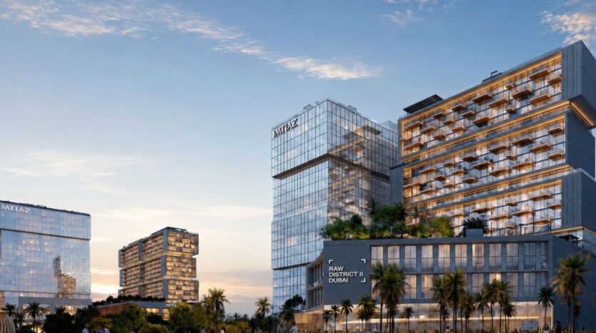 Au coucher du soleil, des bâtiments modernes en verre aux fenêtres illuminées se dressent au milieu des palmiers. Les enseignes « MITAZ » et « Raw District 2 by Imtiaz » ornent le bâtiment principal, se découpant sur un ciel clair aux teintes chaudes.
