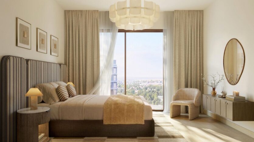 Une chambre moderne de l'établissement « The Canopies by Aldar » comprend un grand lit, une décoration beige, un miroir rond, une fenêtre offrant une vue sur la ville, des rideaux légers, une chaise en bois, des tableaux encadrés et une suspension.