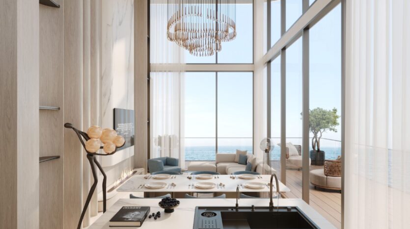 Un espace salon-salle à manger ouvert, moderne et baigné de lumière, avec de hauts plafonds, un lustre, de grandes baies vitrées donnant sur l'océan et un îlot de cuisine élégant au premier plan : découvrez l'élégance raffinée du littoral à Vue Dorée.