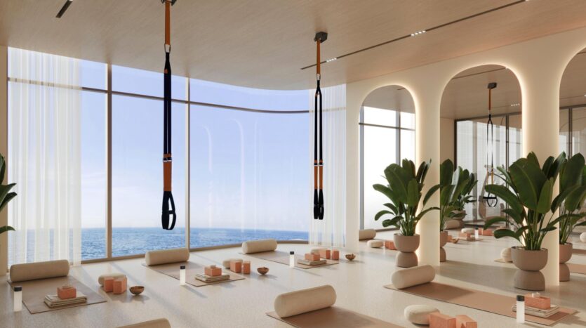Un studio de yoga spacieux et moderne, doté de baies vitrées donnant sur l'océan, situé à Vue Dorée, avec des plantes en pot, des tapis, des coussins de yoga, des blocs de yoga, des bouteilles d'eau et deux sangles de suspension accrochées au plafond.