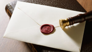 Une enveloppe scellée de couleur crème, ornée d’un cachet de cire rouge apposé à l’aide d’un tampon en laiton, repose sur un livre marron fermé, posé sur une surface en bois : un geste élégant, idéal pour inviter ses proches ou pour envoyer une invitation sophistiquée à Dubaï mettant en avant les atouts de cette ville.