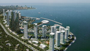 Vue aérienne d'une ville côtière moderne, avec ses gratte-ciel, ses espaces verts et son complexe circulaire en bord de mer, bordé par une mer bleue et calme. On aperçoit des bateaux sur l'eau, qui s'inscrivent dans le décor animé imaginé par HOLM Development, tandis qu'à l'horizon, la ligne d'horizon s'étend le long de la côte.