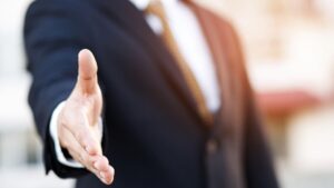 Une personne vêtue d'un costume sombre et d'une cravate tend la main droite pour serrer celle de son interlocuteur, laissant présager le début d'une réunion d'affaires importante — peut-être pour discuter de transactions en difficulté à Dubaï ou pour saisir une aubaine immobilière. L'arrière-plan est légèrement flou, ce qui met l'accent sur l'aspect professionnel des négociations immobilières et du marketing immobilier.