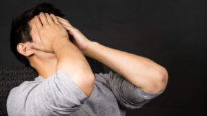 Sur un fond sombre, une personne vêtue d'un t-shirt gris est assise, la tête penchée en arrière et les mains sur le visage, exprimant ainsi la frustration ou le stress que l'on ressent souvent après avoir été confronté aux erreurs commises par les investisseurs immobiliers à Dubaï.
