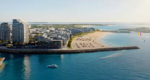 Vue aérienne d'un complexe touristique moderne en bord de mer, avec des immeubles de grande hauteur, une plage de sable, des palmiers, des transats et des personnes. Une eau bleue cristalline, avec un bateau et des kitesurfeurs à proximité.