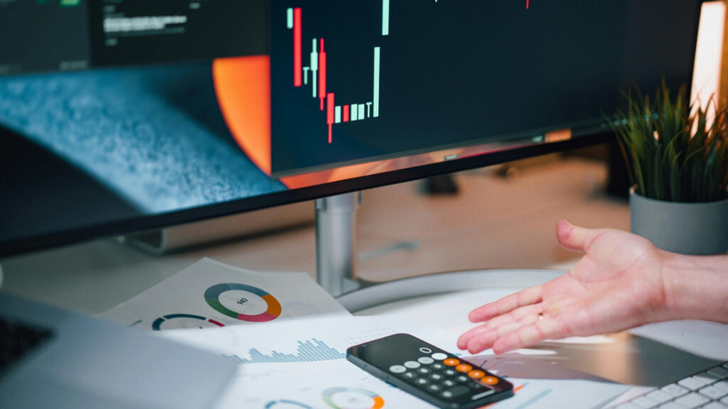 Une main fait un geste près d'un bureau sur lequel se trouvent des graphiques, une calculatrice et des documents, tandis qu'un écran d'ordinateur affiche un graphique boursier en chandeliers rouges et verts — illustrant ainsi le processus d'analyse qui pourrait poser la question suivante : « Est-ce qu'un promoteur immobilier de Dubaï peut faire faillite ? ».