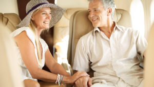 Un couple d'âge mûr, souriant, se tenant la main et assis côte à côte dans les sièges de première classe d'un avion, vêtu de tenues d'été légères et coiffé d'un chapeau de soleil, profitant pleinement de son vol — l'air aussi détendu et raffiné que ce que font différemment les investisseurs les plus riches à Dubaï.