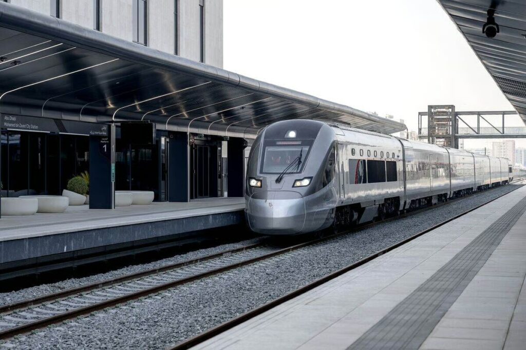 Un train à grande vitesse Etihad Rail moderne, de couleur argentée, est à l'arrêt sur un quai couvert aux lignes épurées, dans une gare contemporaine d'Abou Dhabi aux éléments de design minimalistes.