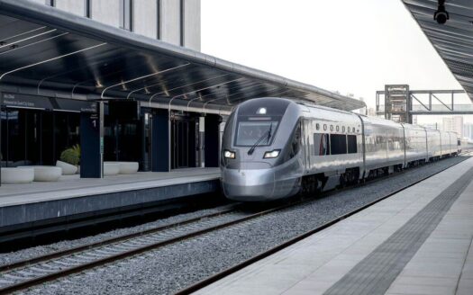 Un train à grande vitesse Etihad Rail moderne, de couleur argentée, est à l'arrêt sur un quai couvert aux lignes épurées, dans une gare contemporaine d'Abou Dhabi aux éléments de design minimalistes.